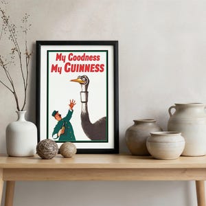 May include: A framed vintage advertisement with the text "My Goodness My Guinness." The artwork features a man in a green coat and hat gesturing towards a large ostrich. The poster is displayed on a wooden table.