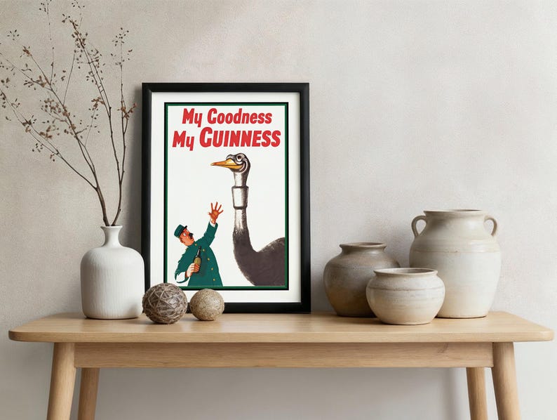 May include: A framed vintage advertisement with the text "My Goodness My Guinness." The artwork features a man in a green coat and hat gesturing towards a large ostrich. The poster is displayed on a wooden table.