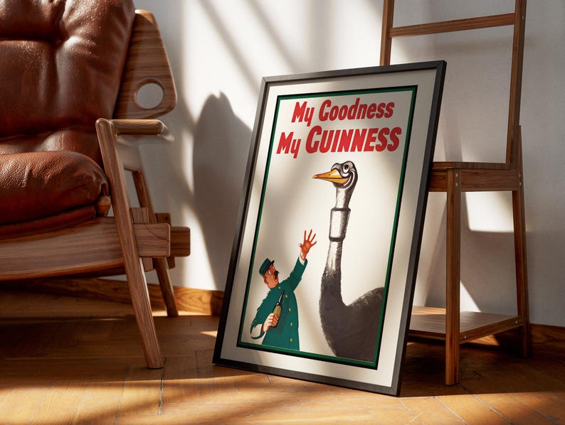 May include: Framed vintage Guinness advertisement featuring a cartoon ostrich with a smiling face and a man in a green uniform. The text "My Goodness My GUINNESS" is displayed in red. The poster is leaning against a wall near a wooden chair.