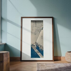 May include: A framed Japanese woodblock print depicting a snowy landscape with a figure on a bridge. The artwork features a dark brown frame, a white mat, and a detailed scene in shades of blue, gray, and white.