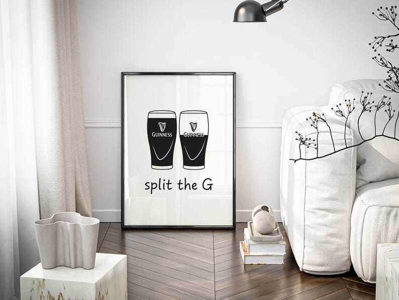 May include: Black and white framed print featuring two Guinness pint glasses. The text "split the G" is below the glasses. The print is displayed in a modern living room setting with a white sofa and decorative elements.