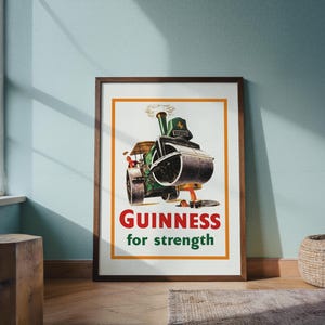 May include: Framed vintage Guinness advert showing a green steamroller with the text "Guinness for strength" in red and green. The artwork has an orange border and a brown wooden frame.
