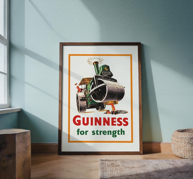 May include: Framed vintage Guinness advert showing a green steamroller with the text "Guinness for strength" in red and green. The artwork has an orange border and a brown wooden frame.