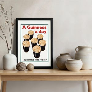 May include: Framed print featuring a vintage advertisement for Guinness. The artwork displays seven pints of Guinness with creamy foam, and the text "A Guinness a day" and "Guinness is good for you" in red and black.