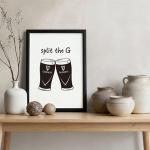 May include: A framed print featuring two Guinness pint glasses with the text "split the G". The glasses are black with white accents and the Guinness logo. The print is displayed on a wooden table with decorative vases.