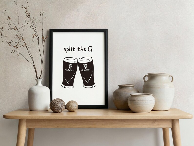 May include: A framed print featuring two Guinness pint glasses with the text "split the G". The glasses are black with white accents and the Guinness logo. The print is displayed on a wooden table with decorative vases.