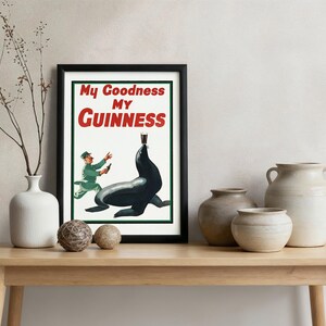 May include: Framed vintage Guinness advertisement featuring a seal balancing a pint of stout. The poster has the text "My Goodness My Guinness" in red and white. The framed poster is displayed on a wooden table with decorative vases and spheres.