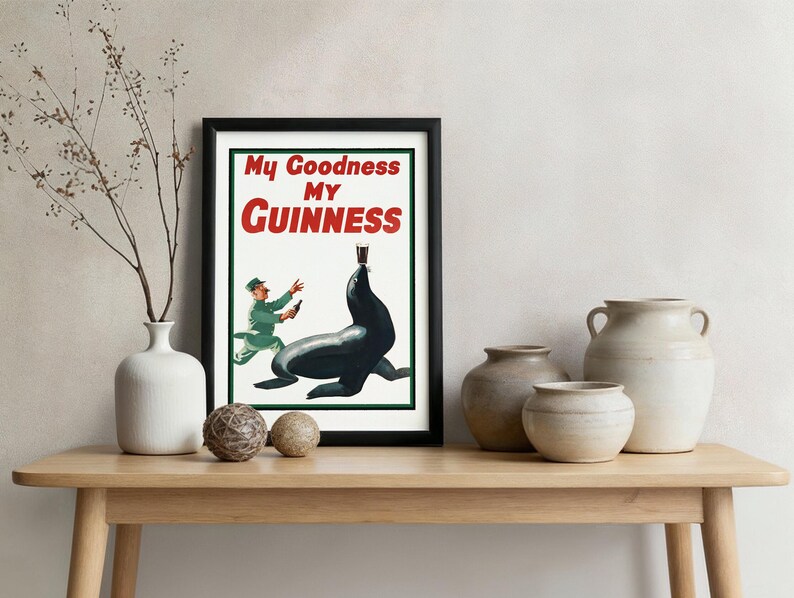 May include: Framed vintage Guinness advertisement featuring a seal balancing a pint of stout. The poster has the text "My Goodness My Guinness" in red and white. The framed poster is displayed on a wooden table with decorative vases and spheres.