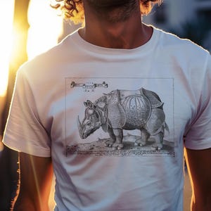 Könnte beinhalten: Weißes T-Shirt mit einer detaillierten Schwarz-Weiß-Illustration eines Nashorns mit Sattel. Die Grafik ist eine Zeichnung im Vintage-Stil, die die Form und Textur des Tieres zeigt. Das Shirt hat einen klassischen Rundhalsausschnitt.