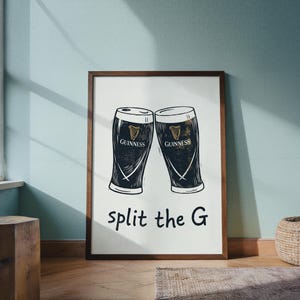 May include: A framed print featuring two Guinness pint glasses, rendered in black and white. The Guinness logo is visible on each glass. The phrase "split the G" is written below the glasses in a simple font. The frame is a dark brown color.