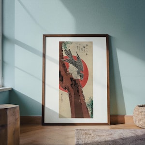 May include: Framed art print featuring a Japanese woodblock print. The artwork depicts a bird of prey perched on a tree trunk, with a red sun in the background. The print has a natural wood frame.