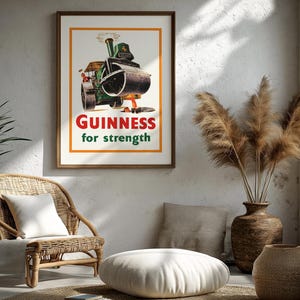 May include: A framed vintage Guinness advert showing a steamroller with the text "Guinness for strength". The artwork is in a wooden frame, displayed in a room with a wicker chair, decorative vases, and a window. The room has a neutral colour scheme.