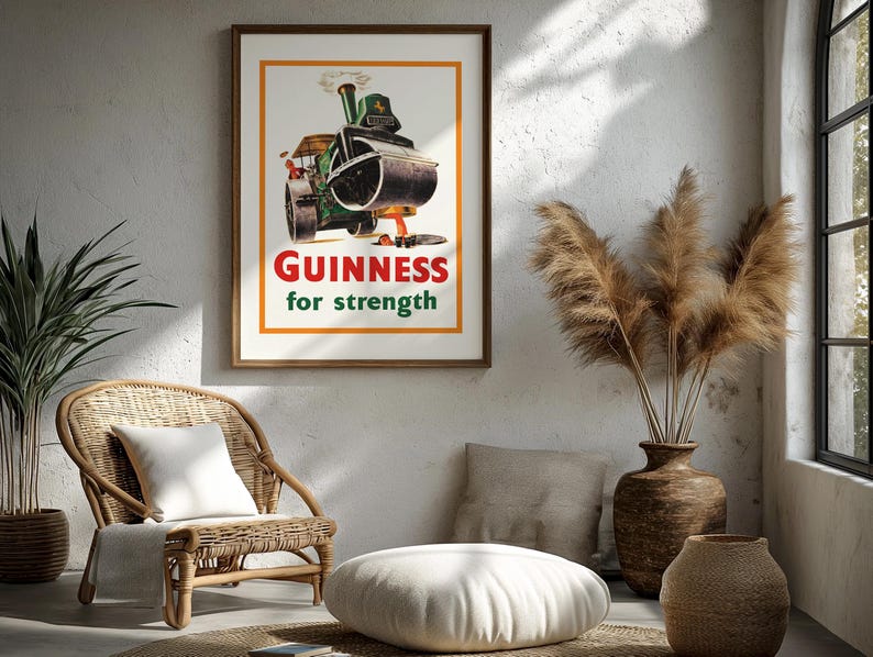 May include: A framed vintage Guinness advert showing a steamroller with the text "Guinness for strength". The artwork is in a wooden frame, displayed in a room with a wicker chair, decorative vases, and a window. The room has a neutral colour scheme.