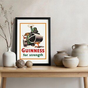 May include: A framed vintage Guinness advert showing a green steamroller with the text "Guinness for strength". The print is in a black frame, displayed on a wooden table with decorative vases and spheres. The image is in a room setting.