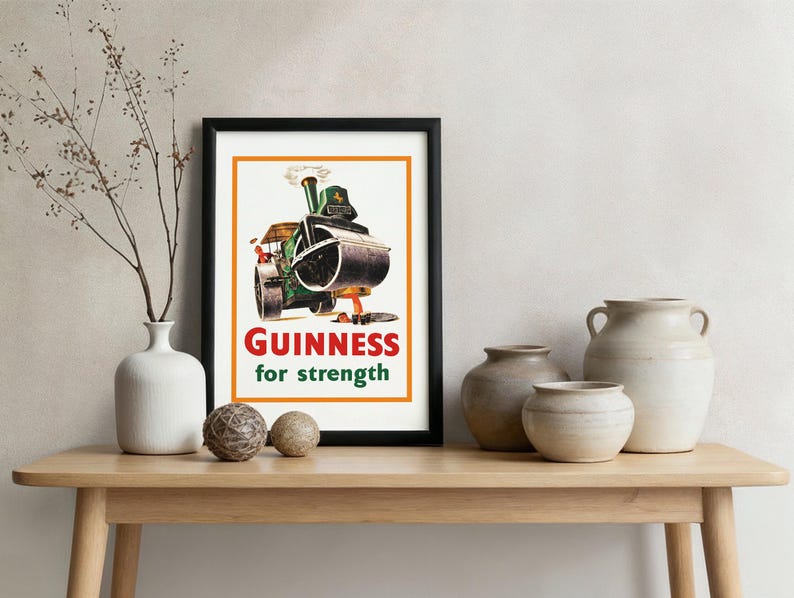 May include: A framed vintage Guinness advert showing a green steamroller with the text "Guinness for strength". The print is in a black frame, displayed on a wooden table with decorative vases and spheres. The image is in a room setting.
