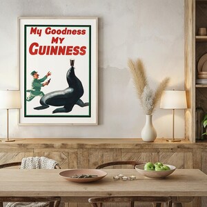 May include: Framed vintage Guinness advertisement featuring a seal balancing a pint of stout. The poster has the text "My Goodness My Guinness" in red and white. A man in a green uniform is running towards the seal. The poster is in a light wood frame.