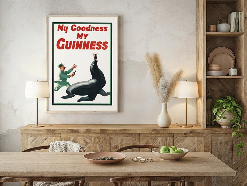 May include: Framed vintage Guinness advertisement featuring a seal balancing a pint of stout. The poster has the text "My Goodness My Guinness" in red and white. A man in a green uniform is running towards the seal. The poster is in a light wood frame.