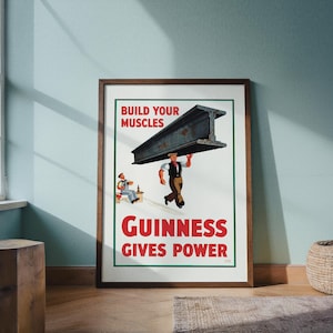 May include: A framed vintage Guinness advertisement. The poster features a man carrying a large steel beam with the text "BUILD YOUR MUSCLES" above and "GUINNESS GIVES POWER" below. A second man is seated, drinking.