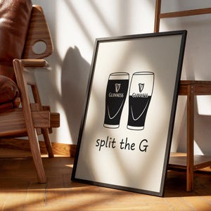 May include: Black and white framed print featuring two Guinness pint glasses with the text "split the G". The print is leaning against a wall, with a wooden chair and shelf in the background.