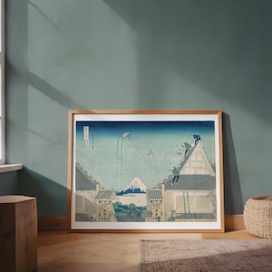 May include: A framed print of a Japanese woodblock print. The artwork depicts a mountain landscape with buildings, people on rooftops, and a kite in the sky. The color palette includes blues, greens, and browns.