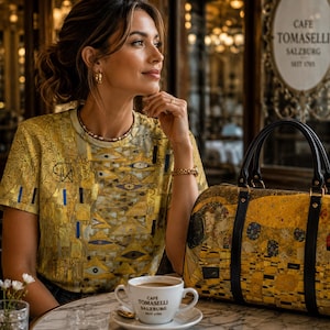 Könnte beinhalten: Ein kurzärmeliges T-Shirt und eine passende Handtasche, beide mit einem goldfarbenen Muster aus geometrischen Formen und Augenmotiven. Eine weiße Keramik-Kaffeetasse mit dem Text "CAFE TOMASELLI SALZBURG SEIT 1705" steht im Vordergrund.