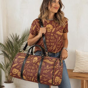 May include: A duffel bag and matching t-shirt in a deep maroon color with a repeating gold pattern of skulls, open books, feathers, and theater masks. The text "TO BE, OR NOT TO BE" is printed in gold on both the bag and the shirt, featuring black straps.