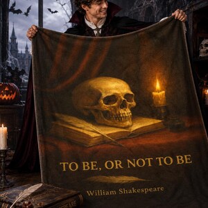 May include: A rectangular throw blanket featuring a dark, atmospheric print of a human skull resting on an open book next to a lit candle. The text "TO BE, OR NOT TO BE" and "William Shakespeare" is printed in gold lettering on the lower section.