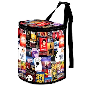 Puede incluir: Una bolsa nevera cilíndrica negra cubierta de coloridos carteles de teatro musical. La bolsa tiene una cremallera negra, una correa para el hombro negra y una parte superior negra. Los carteles incluyen títulos como "Wicked", "Mamma Mia" y "Chicago".