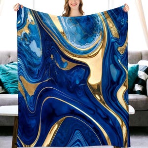 May include: A blue and gold abstract patterned blanket. The blanket features a swirling design with shades of blue and gold. The gold accents create a luxurious and elegant look. The blanket is held up by a person.