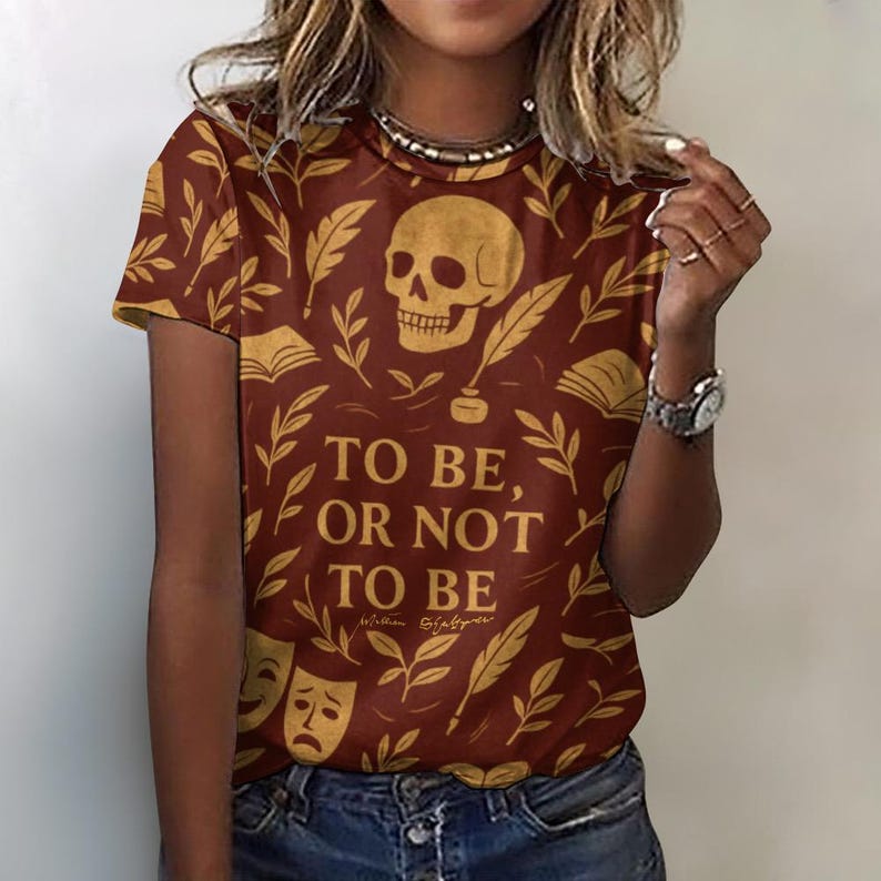 Maroon t-shirt with a repeating pattern of gold-colored leaves, books, and a skull. The text "TO BE, OR NOT TO BE" is displayed in a bold font, with "William Shakespeare" below. HALLOWEEN shirt with skull, spooky christmass shirt