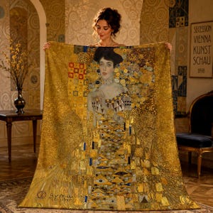 May include: A rectangular throw blanket featuring a reproduction of Gustav Klimt's Portrait of Adele Bloch-Bauer I. The design displays a woman in a gold-toned dress with intricate geometric patterns, swirling motifs, and eye-shaped symbols.