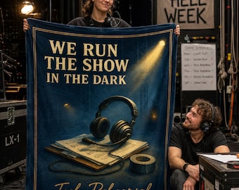 May include: A dark blue rectangular fleece blanket featuring the gold text "WE RUN THE SHOW IN THE DARK" at the top and "Tech Rehearsal" at the bottom. The center graphic depicts a pair of black headphones resting on a stack of papers and a roll of tape.