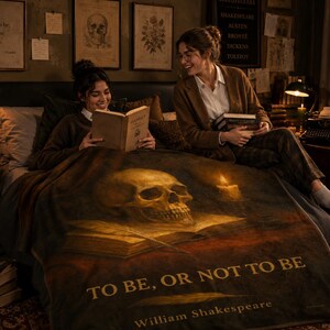 May include: A dark-toned throw blanket featuring a large human skull resting on an open book, a lit candle, and the text "TO BE, OR NOT TO BE" followed by "William Shakespeare" in gold lettering, displayed over a bed in a dimly lit room.