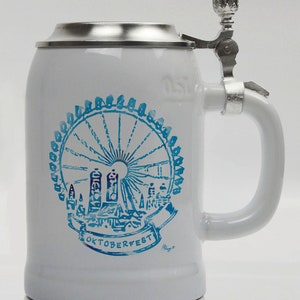 Op de afbeelding: Witte keramische bierpul met een blauw reuzenradontwerp en het woord "Oktoberfest". De pul heeft een metalen scharnierend deksel en handvat. De pul is ongeveer 18 cm hoog.