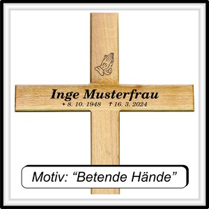 May include: A wooden cross with a black inscription that reads "Inge Musterfrau * 8. 10. 1948 &dagger; 16. 3. 2024". The cross also features a black silhouette of praying hands. A smaller plaque below reads "Motiv: "Betende H&auml;nde"".
