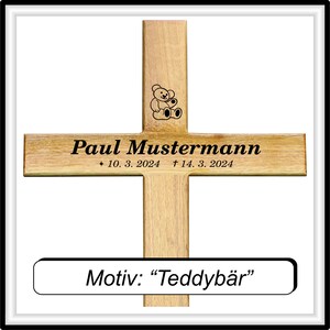 May include: A wooden cross with a teddy bear design engraved on the top. The cross is inscribed with the name "Paul Mustermann" and the dates "* 10. 3. 2024 &dagger; 14. 3. 2024". Below the cross is a small plaque that reads "Motiv: "Teddyb&auml;r"".