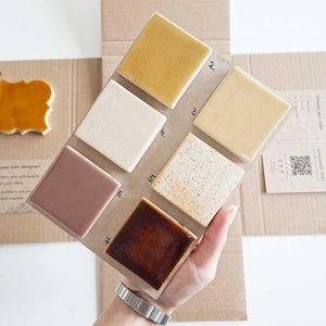 May include: A sample board of handmade tiles in various colors, including beige, brown, and off-white. The tiles are arranged on a cardboard backing, showcasing different textures and finishes. The board is held by a person.