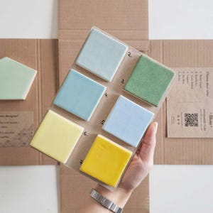 Può includere: Una cartella colori con sei piastrelle quadrate nei toni verde pastello, eucalipto, patina verde, laguna, azzurro cielo e giallo burro, disposte su due colonne. Una piastrella pentagonale è visibile a sinistra, con testo stampato e un QR code a destra.