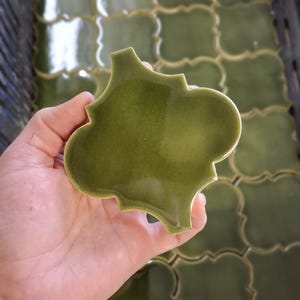 Puede incluir: Un azulejo de cerámica de color verde oliva con forma de cuatrifolio y acabado brillante, sostenido para mostrar su superficie lisa y bordes curvos. El azulejo presenta un color verde uniforme y un diseño simétrico de cuatro lóbulos.