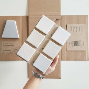 Può includere: Una selezione di campioni di piastrelle fatte a mano in varie tonalità di bianco e grigio, esposti su un supporto di cartone. I campioni includono piastrelle quadrate e una forma trapezoidale unica. Il testo "Be your own designer!" è visibile.