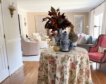 May include: A round table covered with a floral tablecloth featuring pink and green flowers, with a fringed edge. The table is decorated with blue and white vases, a small pumpkin, and a large arrangement of red leaves. A pink armchair is visible in the background.