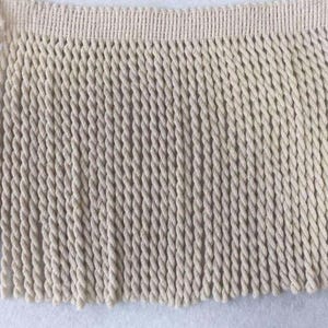 May include: Close-up of a cream-coloured fringe trim. The trim features a woven top section with a series of tightly twisted rope-like strands hanging below. The strands are uniform in length, creating a textured, decorative element.