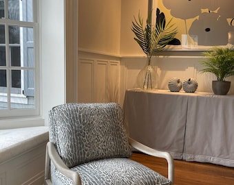 May include: An upholstered armchair with a gray and white animal print pattern. The chair has a light wood frame and is placed in a room with a large abstract floral painting and a window with white shutters.