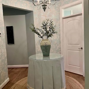 Peut inclure: Une jupe de table bleu clair à plis, un plateau de table en verre et un vase en céramique blanche avec un motif floral vert. Le vase est rempli d'une plante verte avec des feuilles vert foncé et de petites baies noires. La table est dans un couloir avec une porte blanche et un papier peint à motifs bleu clair.