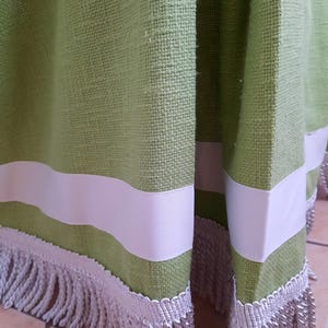 Pode incluir: Close-up de um tecido verde com uma faixa branca e franjas prateadas. O tecido tem uma textura tecida. A faixa branca é uma fita larga. A franja é uma corda grossa prateada.