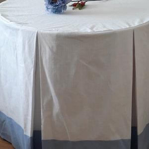 May include: A white round tablecloth with a blue trim. The tablecloth has pleats around the bottom edge. A single blue flower with a green stem is lying on the tablecloth.