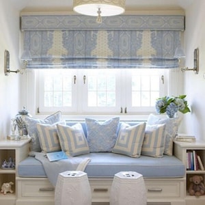May include: A white window seat with blue cushions and pillows, a white patterned valance, and two white ceramic stools. The window seat is built into a white cabinet with shelves.