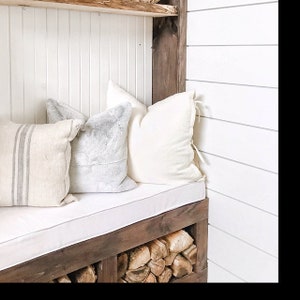 May include: A white cushioned bench with three pillows, one striped, one gray, and one white, sits in front of a wooden shelf with stacked firewood underneath. The wall behind the bench is white with horizontal wood paneling.