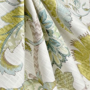 May include: A white fabric with a floral pattern in shades of green, blue, and brown. The fabric has a textured, linen-like appearance.