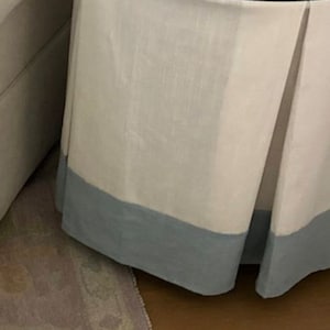 Op de afbeelding: Een ronde tafel met een witte stoffen rok en een blauwe zoom. De tafel heeft een glazen blad.